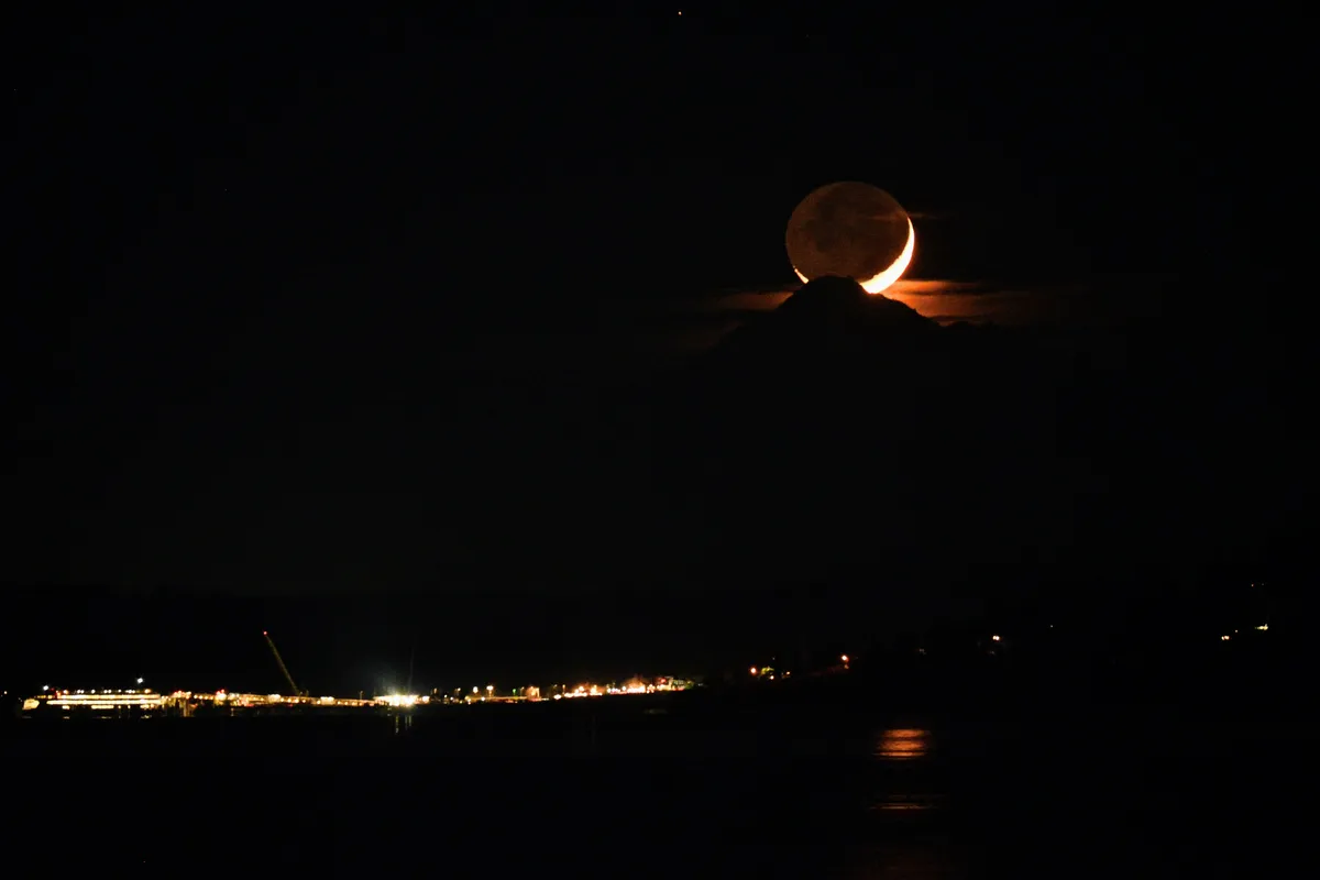 A crescent moon, the rest of its disc faintly lit, sits atop a dark mountain above Kingston's lights across Puget Sound.