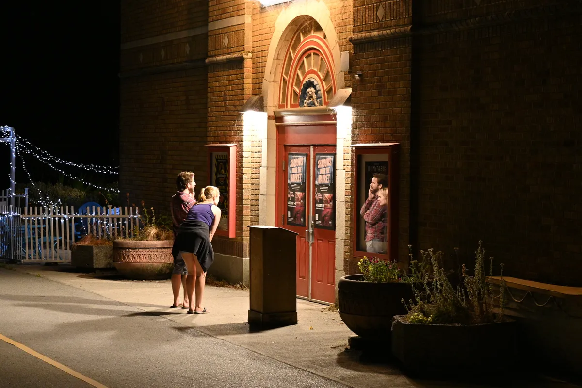 A bearded man in plaid and a blonde woman read a theater poster at night; across the doorway, a display case's glass reflects them.